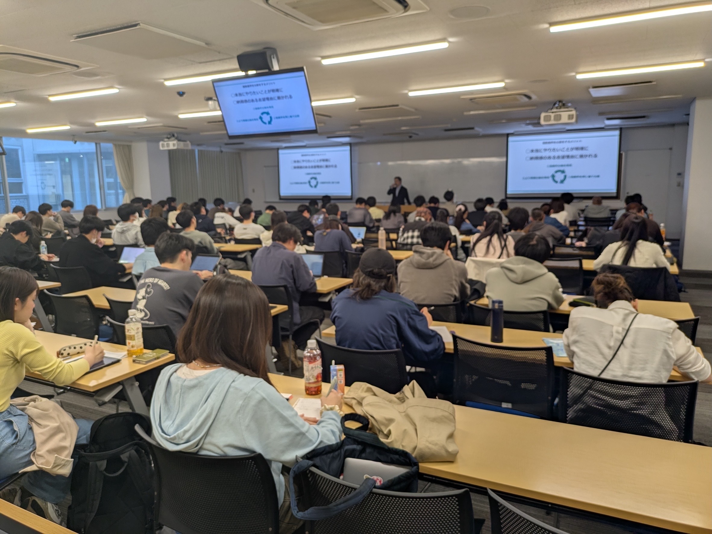 大学の教室で、登壇者の説明を聞く学生たちと講義スクリーン